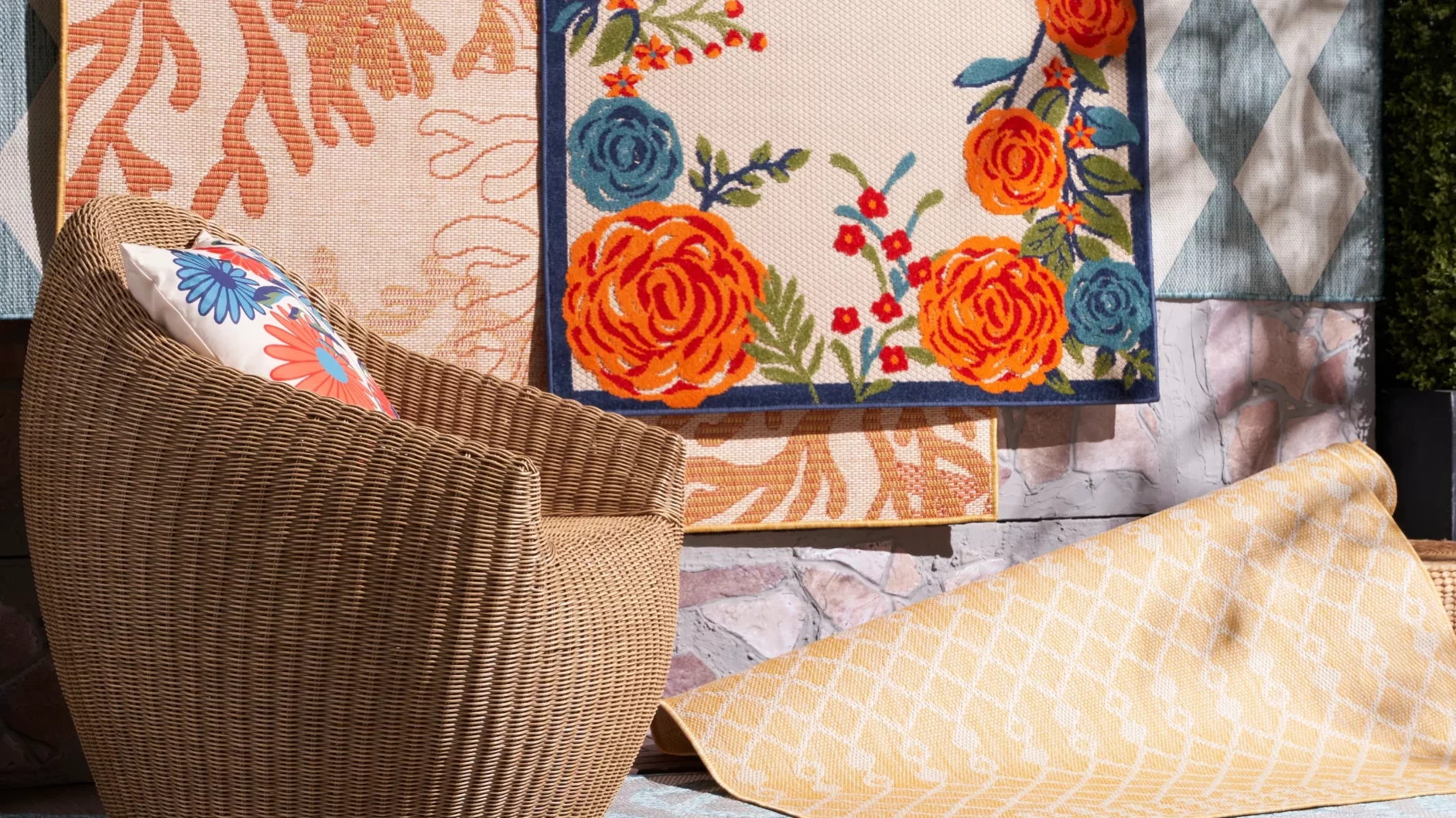 Nourison Positano outdoor rugs in bold floral and geometric patterns displayed against a dark hedge backdrop with a wicker chair in the foreground
